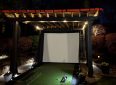 Outdoor projector screen under a wooden pergola at night with landscape lighting.